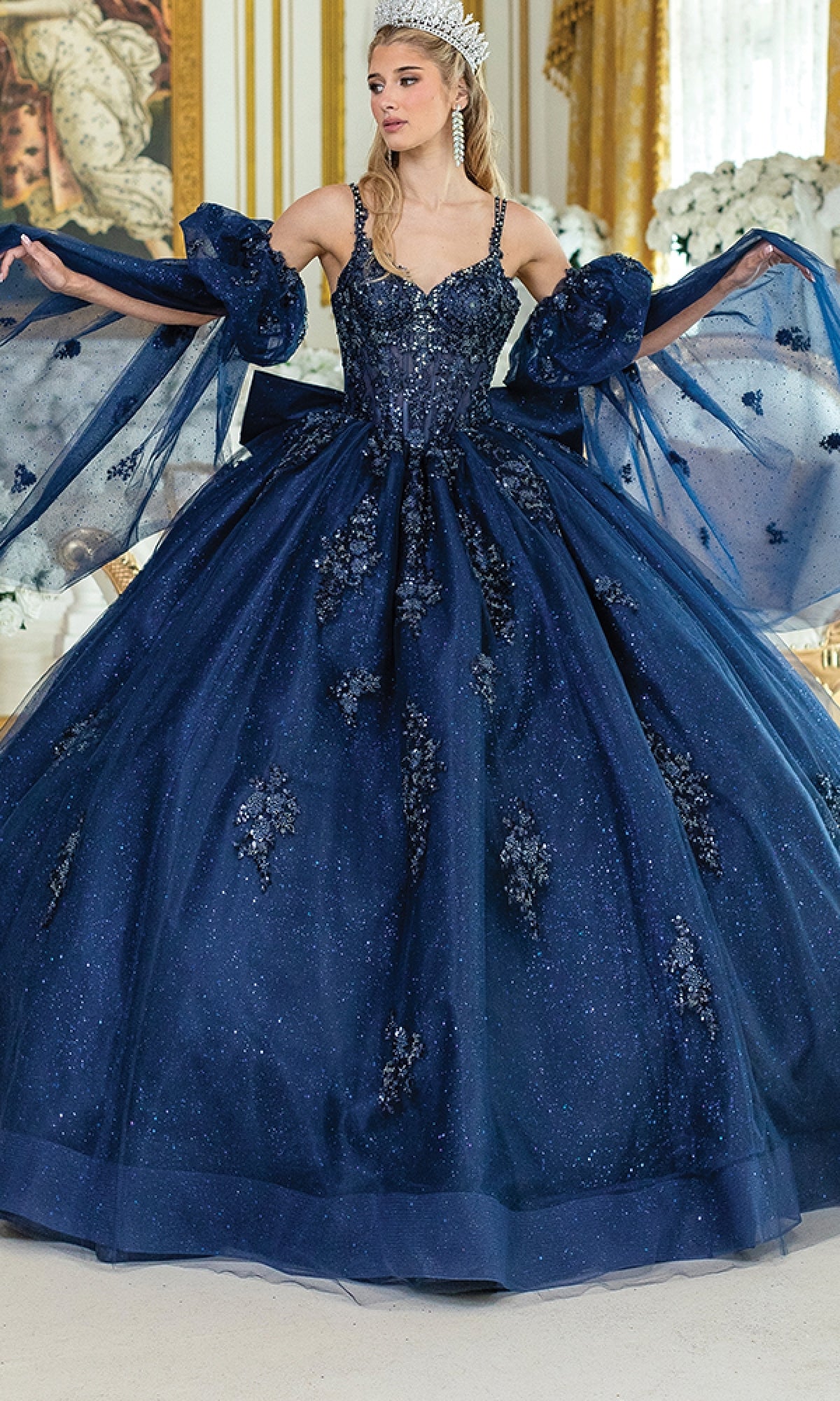 A woman stands indoors wearing the Blue Quinceanera Dress 5037 By Dancing Queen, a sparkling navy ball gown with floral details and puffed sleeves, holding her skirt.